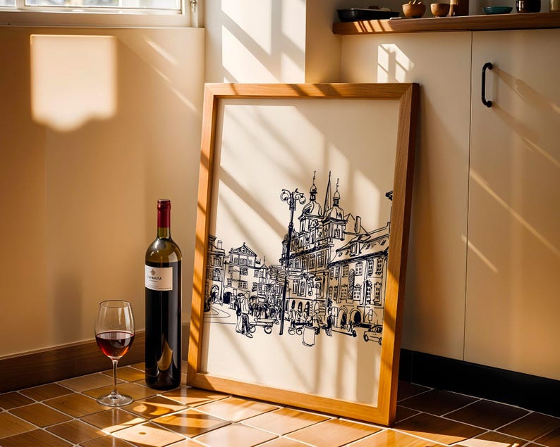 May include: A framed print of a European city scene in black and white, leaning against a wall. A bottle of red wine and a wine glass with red wine are in front of the frame. The frame is made of light brown wood.