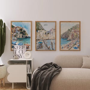 May include: Three framed watercolor prints of coastal Italian towns with colorful buildings and blue water. The prints are in wooden frames and hang above a beige sofa. A tall cactus in a white pot sits to the left.