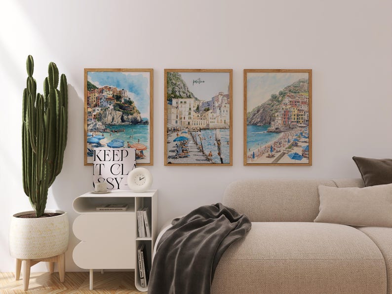 May include: Three framed watercolor prints of coastal Italian towns with colorful buildings and blue water. The prints are in wooden frames and hang above a beige sofa. A tall cactus in a white pot sits to the left.