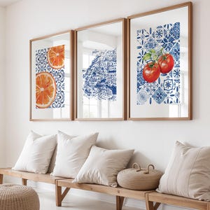 May include: Three framed art prints with wooden frames. The prints feature watercolor illustrations of an orange slice, a coastal town, and tomatoes. The backgrounds have a blue and white tile pattern. The prints are displayed above a wooden bench with pillows.