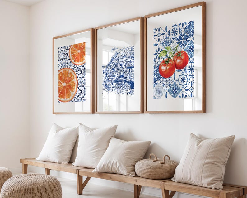 May include: Three framed art prints with wooden frames. The prints feature watercolor illustrations of an orange slice, a coastal town, and tomatoes. The backgrounds have a blue and white tile pattern. The prints are displayed above a wooden bench with pillows.
