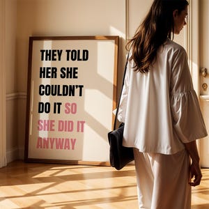 May include: A framed print with the text "THEY TOLD HER SHE COULDN'T DO IT SO SHE DID IT ANYWAY" in black and pink. A person wearing a white outfit and carrying a black bag stands near the print.