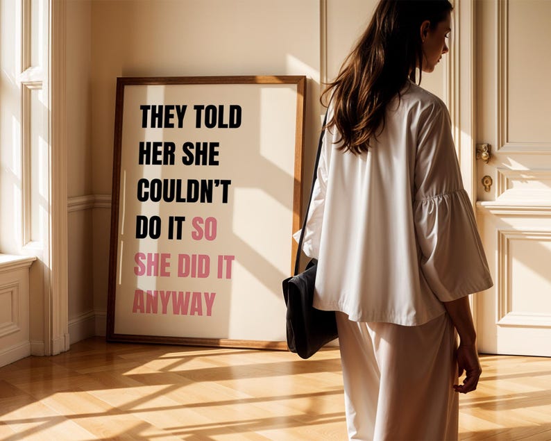 May include: A framed print with the text "THEY TOLD HER SHE COULDN'T DO IT SO SHE DID IT ANYWAY" in black and pink. A person wearing a white outfit and carrying a black bag stands near the print.