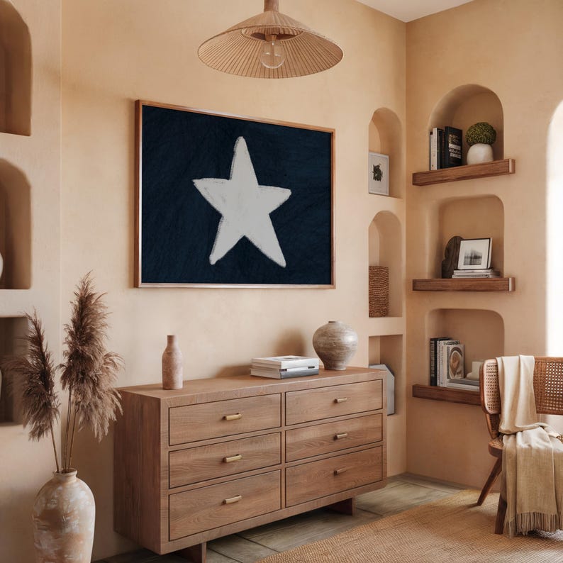 May include: A framed artwork featuring a large white star on a dark blue background hangs above a wooden dresser. The room has a neutral color palette with a woven light fixture and decorative items.