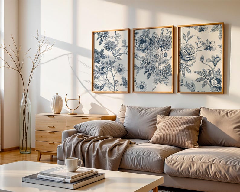 May include: Three framed floral art prints. Each print features a blue and white botanical design. The prints are displayed above a beige sectional sofa. A wooden cabinet, a vase with branches, and a coffee table with books and a mug complete the living room setting.