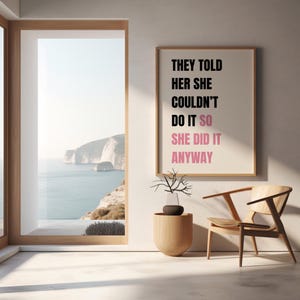 May include: A framed print with the text "THEY TOLD HER SHE COULDN'T DO IT SO SHE DID IT ANYWAY" in black and pink. The print hangs on a white wall next to a wooden chair and a decorative vase. A large window reveals a coastal view.