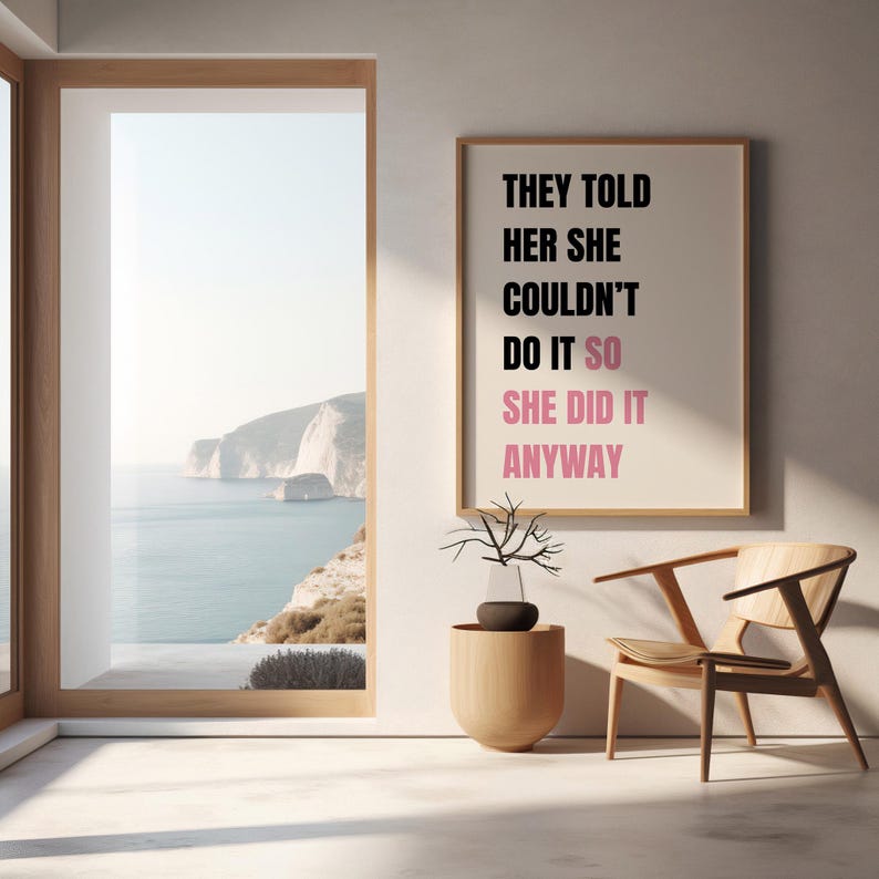 May include: A framed print with the text "THEY TOLD HER SHE COULDN'T DO IT SO SHE DID IT ANYWAY" in black and pink. The print hangs on a white wall next to a wooden chair and a decorative vase. A large window reveals a coastal view.