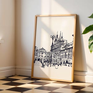 May include: A framed black and white architectural sketch of a European city scene. The artwork features detailed line drawings of buildings, a street lamp, and figures, set against a cream-colored background. The frame is a light wood color, and the artwork is leaning against a wall.