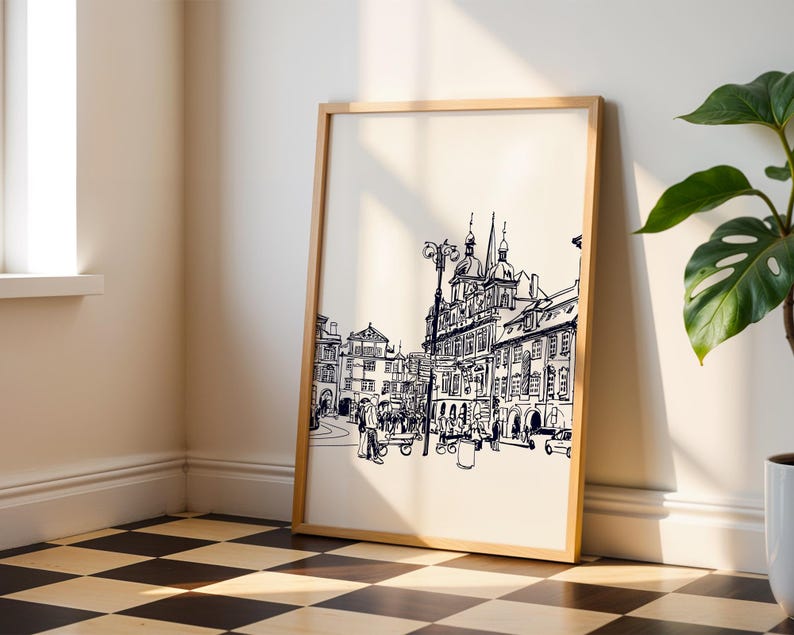 May include: A framed black and white architectural sketch of a European city scene. The artwork features detailed line drawings of buildings, a street lamp, and figures, set against a cream-colored background. The frame is a light wood color, and the artwork is leaning against a wall.
