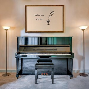 Puede incluir: Un piano vertical negro con un banco a juego se encuentra en una habitaci&oacute;n con dos l&aacute;mparas de pie. Encima del piano hay un grabado enmarcado con las palabras "tell me when" y una ilustraci&oacute;n de vino verti&eacute;ndose.