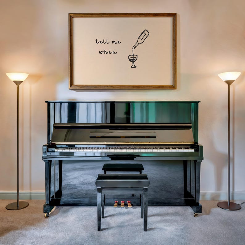 Puede incluir: Un piano vertical negro con un banco a juego se encuentra en una habitaci&oacute;n con dos l&aacute;mparas de pie. Encima del piano hay un grabado enmarcado con las palabras "tell me when" y una ilustraci&oacute;n de vino verti&eacute;ndose.