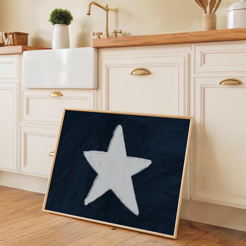May include: A framed art print featuring a large, white star against a dark blue background. The artwork has a textured appearance and is set in a light wood frame. The print is leaning against a kitchen cabinet.