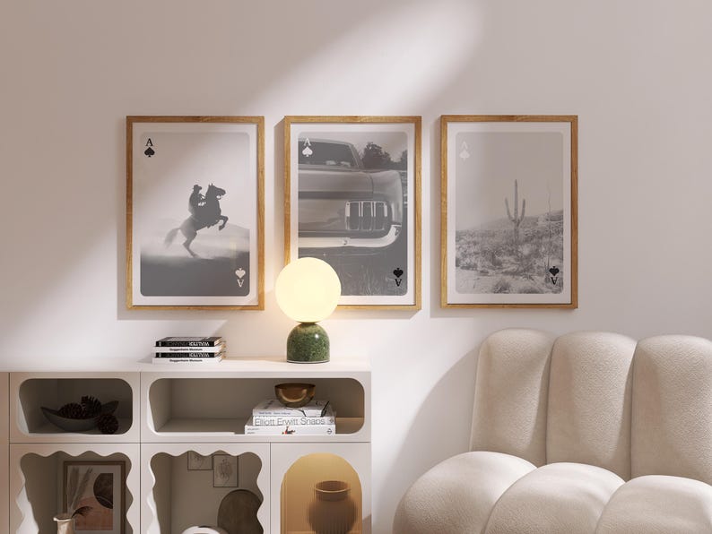 Op de afbeelding: Drie ingelijste kunstdrukken met een speelkaartthema. De prints tonen een cowboy op een paard, een auto en een woestijnlandschap. Een groene en gouden lamp staat op een witte kast.