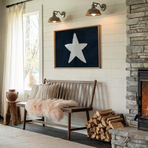 May include: A rustic interior scene featuring a wooden bench with striped pillows and a pink fur throw. A framed artwork with a white star on a navy background hangs above the bench. A fireplace and firewood add warmth.
