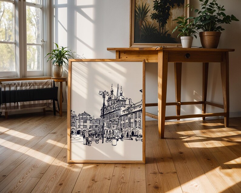 May include: A framed black and white architectural print of a European city scene. The print is in a light wood frame and leans against a wall. The image features detailed line drawings of buildings, streets, and people. The room has hardwood floors and wooden furniture.