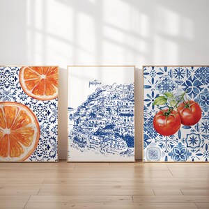 May include: Three framed art prints. One features orange slices on a blue and white patterned background. The second depicts a blue and white illustration of a coastal town. The third shows tomatoes on a blue and white patterned background.