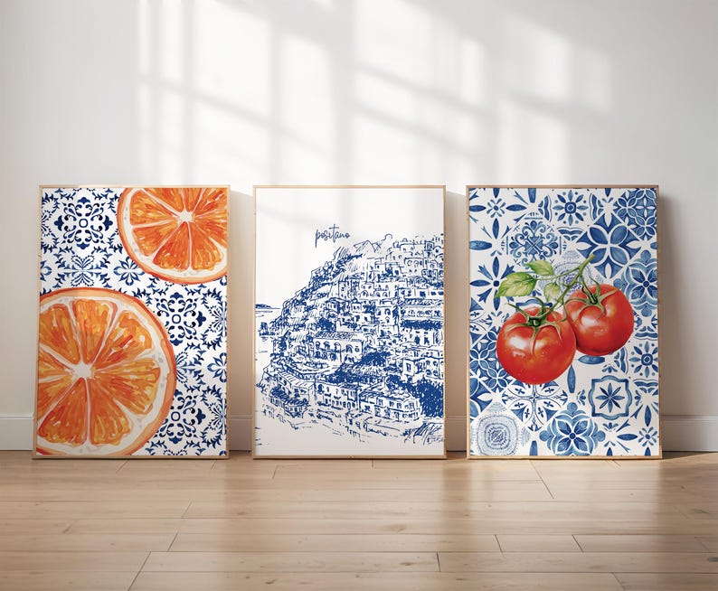 May include: Three framed art prints. One features orange slices on a blue and white patterned background. The second depicts a blue and white illustration of a coastal town. The third shows tomatoes on a blue and white patterned background.