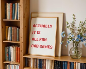 May include: A framed print with the text "ACTUALLY IT IS ALL FUN AND GAMES" in red letters. The print is displayed on a wooden shelf with a bookcase and a vase of blue flowers. The bookcase is filled with books.
