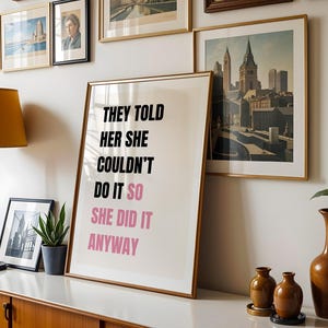 May include: A framed print with the text "THEY TOLD HER SHE COULDN'T DO IT SO SHE DID IT ANYWAY" in black and pink. The print is displayed on a white surface with other framed art, a lamp, and decorative vases. The overall theme is inspirational.