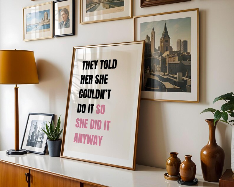 May include: A framed print with the text "THEY TOLD HER SHE COULDN'T DO IT SO SHE DID IT ANYWAY" in black and pink. The print is displayed on a white surface with other framed art, a lamp, and decorative vases. The overall theme is inspirational.