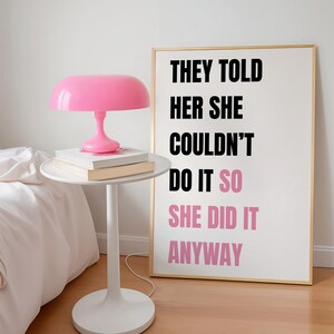May include: A pink mushroom-shaped lamp sits on a white side table with books. A framed print with gold trim displays the text "THEY TOLD HER SHE COULDN'T DO IT SO SHE DID IT ANYWAY" in black and pink.