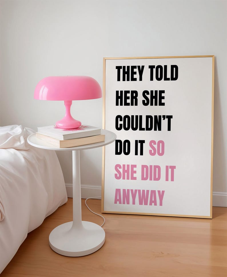 May include: A pink mushroom-shaped lamp sits on a white side table with books. A framed print with gold trim displays the text "THEY TOLD HER SHE COULDN'T DO IT SO SHE DID IT ANYWAY" in black and pink.