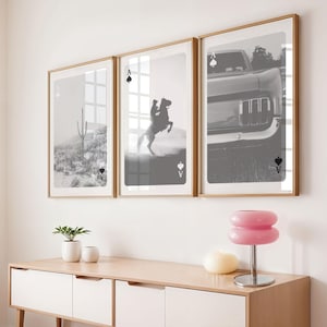 Op de afbeelding: Drie ingelijste zwart-wit prints met speelkaartmotieven. De prints tonen een woestijnlandschap, een cowboy op een steigerend paard en een klassieke auto. De lijsten zijn licht van kleur, boven een houten kast.