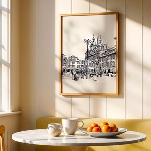 May include: A framed black and white sketch of a European city scene hangs on a cream-colored wall. A round table with a marble top holds a white mug, a small ceramic cup, and a plate of oranges. The scene is illuminated by sunlight.