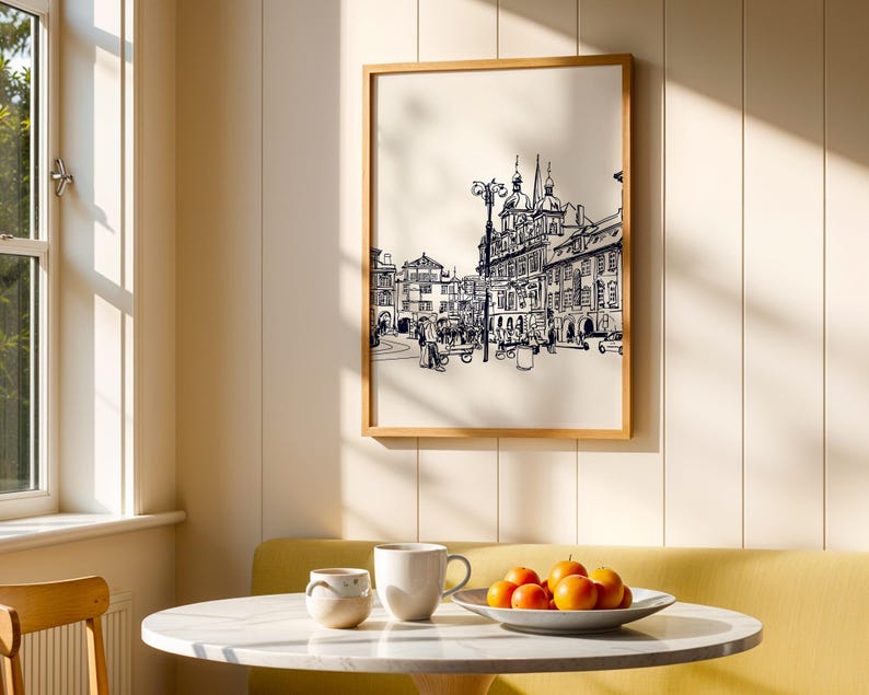 May include: A framed black and white sketch of a European city scene hangs on a cream-colored wall. A round table with a marble top holds a white mug, a small ceramic cup, and a plate of oranges. The scene is illuminated by sunlight.