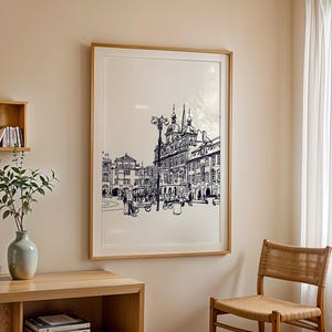 May include: A framed black and white architectural print depicting a European city square. The artwork is in a light wood frame and hangs on a beige wall. A wooden shelf with books and a vase with greenery are in the foreground.