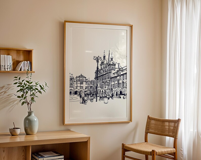 May include: A framed black and white architectural print depicting a European city square. The artwork is in a light wood frame and hangs on a beige wall. A wooden shelf with books and a vase with greenery are in the foreground.