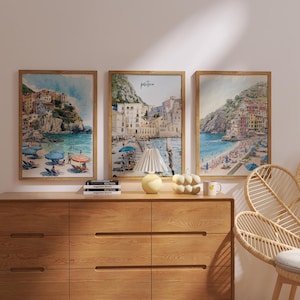 May include: Three framed watercolor prints depicting coastal scenes with colorful buildings, beaches, and umbrellas. The artwork is displayed above a wooden dresser. A decorative lamp, a ceramic pumpkin, and a mug are on the dresser.