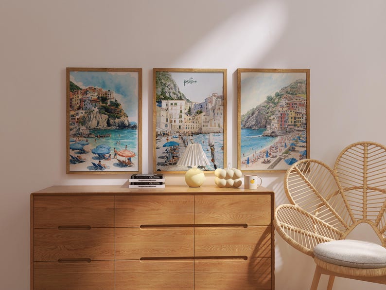 May include: Three framed watercolor prints depicting coastal scenes with colorful buildings, beaches, and umbrellas. The artwork is displayed above a wooden dresser. A decorative lamp, a ceramic pumpkin, and a mug are on the dresser.