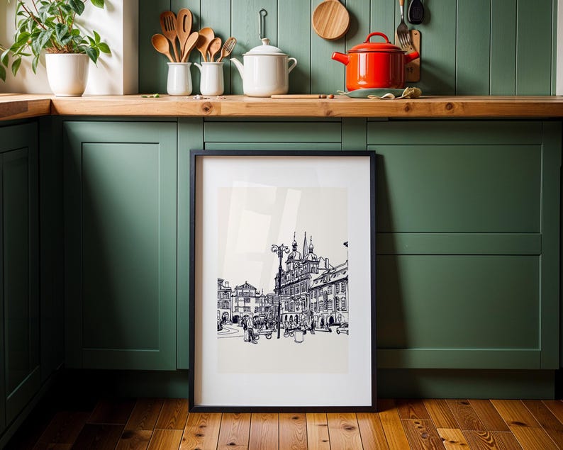 May include: A framed black and white architectural print featuring a detailed street scene with buildings, streetlights, and figures. The artwork is displayed against a neutral background, with a black frame. The print is set against a backdrop of a green kitchen.