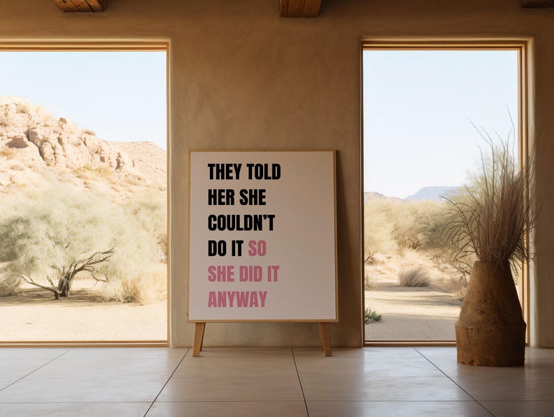 May include: A framed print with the text "THEY TOLD HER SHE COULDN'T DO IT SO SHE DID IT ANYWAY" in black and pink. The print is on a wooden easel in front of a window with a desert landscape.