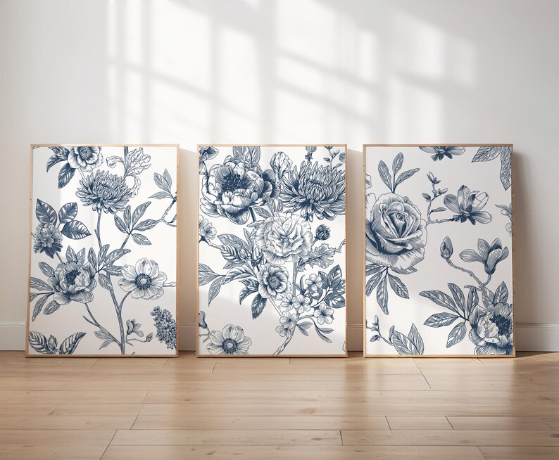 Pu&ograve; includere: Tre stampe artistiche floreali incorniciate. Ogni stampa presenta una diversa disposizione di fiori blu e bianchi, tra cui rose, peonie e altri fiori. L'opera d'arte &egrave; esposta su un pavimento in legno chiaro.