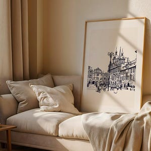 May include: A framed black and white architectural print of a European city scene. The artwork is displayed on a beige wall above a beige sofa with pillows and a blanket. The room has beige curtains and a wooden side table.