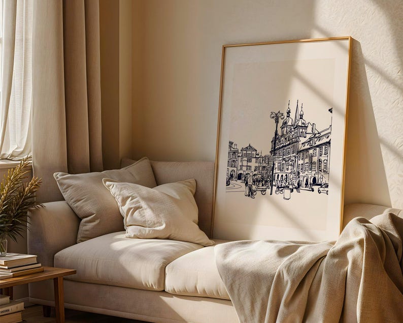 May include: A framed black and white architectural print of a European city scene. The artwork is displayed on a beige wall above a beige sofa with pillows and a blanket. The room has beige curtains and a wooden side table.
