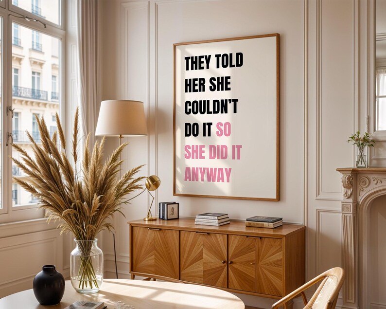 May include: A framed print with the text "THEY TOLD HER SHE COULDN'T DO IT SO SHE DID IT ANYWAY" in black and pink. The print hangs on a white wall above a wooden cabinet. A vase of dried plants sits on a table in front of a window.