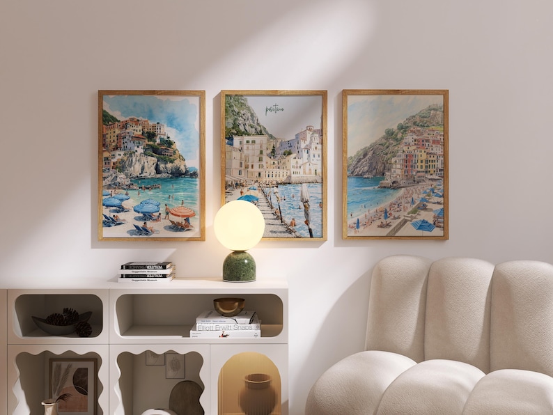 May include: Three framed watercolor prints depict coastal scenes with colorful buildings, beaches, and umbrellas. The artwork is displayed above a white cabinet with a green lamp and a beige sofa. The prints are in wooden frames.