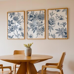 Pu&ograve; includere: Tre stampe floreali incorniciate con disegni botanici blu e bianchi sono appese sopra un tavolo e sedie in legno. Le stampe presentano illustrazioni dettagliate di fiori e fogliame. Un vaso grigio con fiori gialli &egrave; sul tavolo.