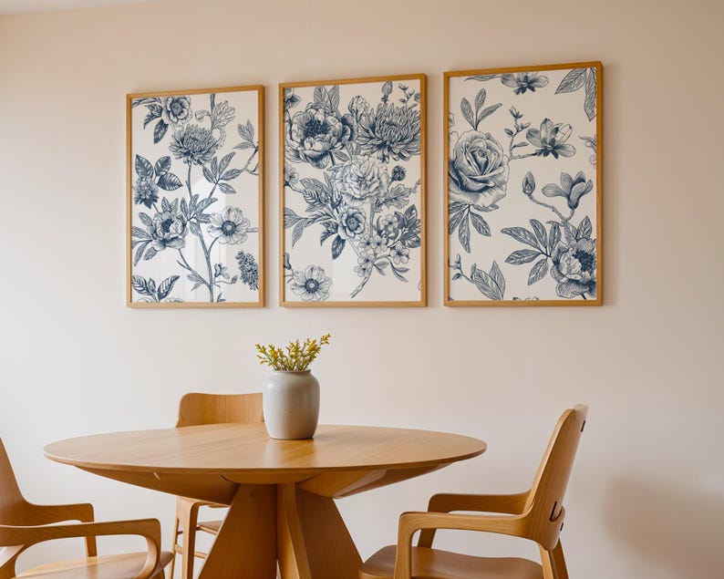 Pu&ograve; includere: Tre stampe floreali incorniciate con disegni botanici blu e bianchi sono appese sopra un tavolo e sedie in legno. Le stampe presentano illustrazioni dettagliate di fiori e fogliame. Un vaso grigio con fiori gialli &egrave; sul tavolo.