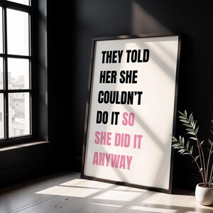 May include: Framed art print with the text "THEY TOLD HER SHE COULDN'T DO IT SO SHE DID IT ANYWAY" in black and pink. The print is in a dark wooden frame, leaning against a black wall. A small potted plant sits to the right.
