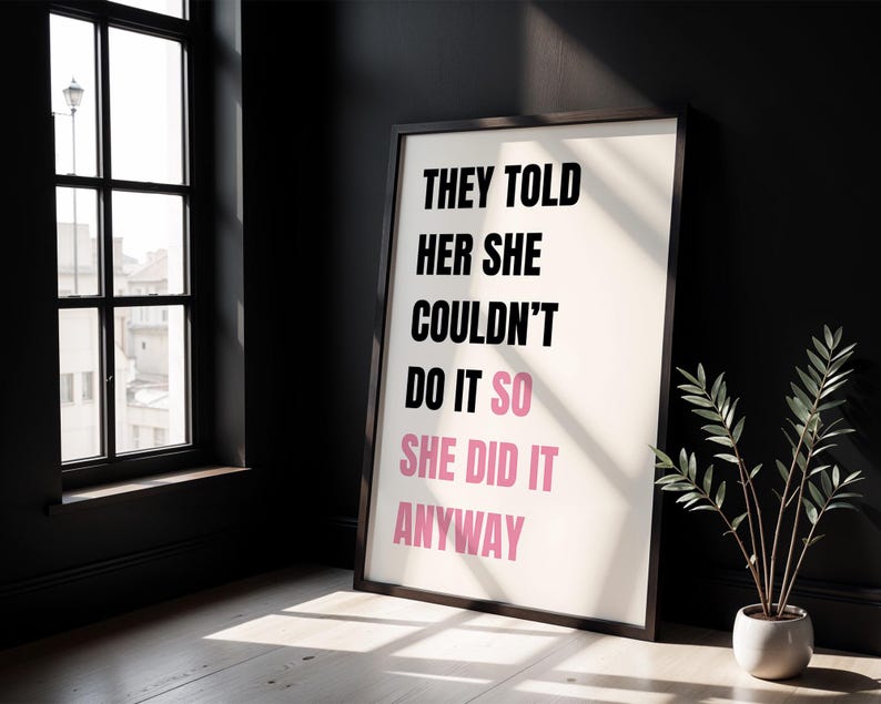 May include: Framed art print with the text "THEY TOLD HER SHE COULDN'T DO IT SO SHE DID IT ANYWAY" in black and pink. The print is in a dark wooden frame, leaning against a black wall. A small potted plant sits to the right.