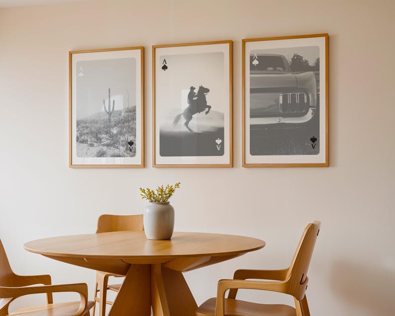 May include: Three framed art prints with playing card designs. The prints feature black and white photographs: a cactus in a desert, a cowboy on a horse, and a car's front. Each print has a playing card symbol: Ace of Spades, Ace of Spades, and Ace of Spades. The prints are displayed above a wooden table with chairs.