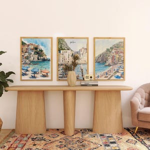 May include: Three framed watercolor prints of coastal scenes with colorful buildings and blue water hang above a wooden console table. A potted plant, vase, and decorative radio are on the table. A patterned rug and a pink sofa complete the room.