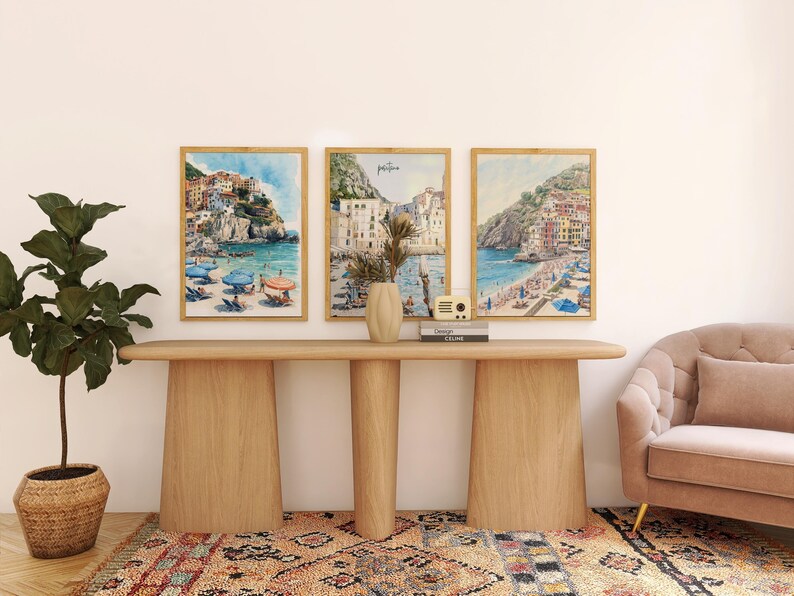 May include: Three framed watercolor prints of coastal scenes with colorful buildings and blue water hang above a wooden console table. A potted plant, vase, and decorative radio are on the table. A patterned rug and a pink sofa complete the room.