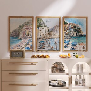 May include: Three framed watercolor prints of coastal scenes with buildings, beaches, and umbrellas. The prints are in wooden frames and are displayed above a white cabinet with gold handles. A small sign and snacks are on the cabinet.