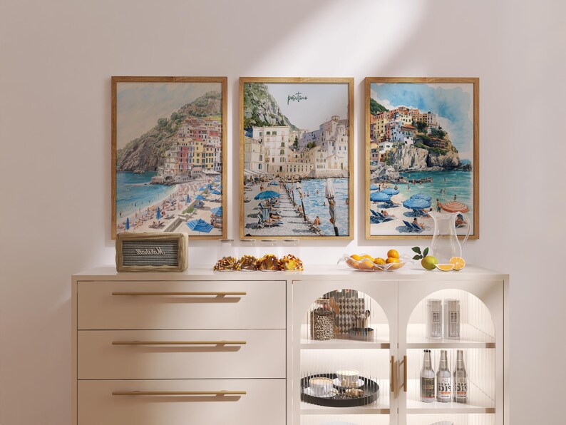 May include: Three framed watercolor prints of coastal scenes with buildings, beaches, and umbrellas. The prints are in wooden frames and are displayed above a white cabinet with gold handles. A small sign and snacks are on the cabinet.