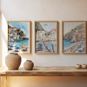 May include: Three framed watercolor prints of coastal scenes with buildings and beaches. The prints are in light wood frames. Two beige ceramic vases and three small beige ceramic bowls sit on a wooden table. The window frame is visible on the left.